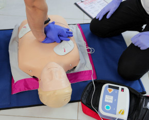 handen doen hartmassage op oefenpop naast een AED toestel